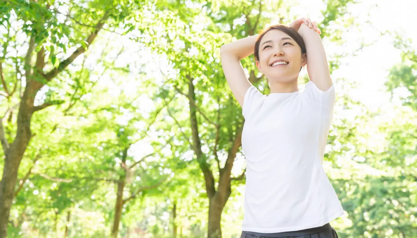 緑あふれる公園で、白いTシャツを着た女性が両腕を伸ばして笑顔でストレッチをしている